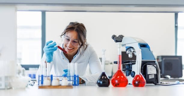 Scientist wearing a lab coat and protective glasses smiles while examining a test tube containing a red liquid in a laboratory setting, surrounded by other lab equipment